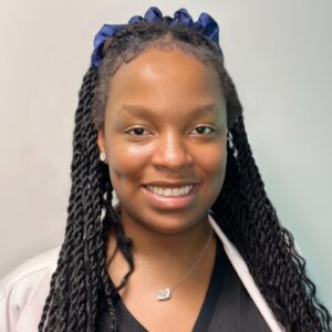 A headshot of Dr. Wynette Bender in front of a white wall. She is wearing black scrubs, a white lab coat, jewelry, and a blue ribbon in her hair.