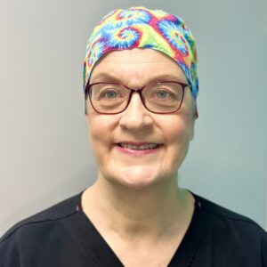 A headshot of Dr. Andrea Miller in front of a white background. She is wearing black scrubs, glasses, and a rainbow swirl hat.